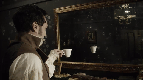 A man in old fashioned clothing stands in front of a mirror holding a tea cup. In the mirror, only the tea cup is visible. 